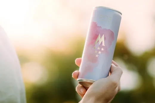 Eine Hand hält eine pastellfarbene Dose mit goldener Beschriftung und einem rosa Aquarellmotiv. Auf der Dose ist ein großes „M“ mit Sternen darunter sowie das Wort „Rosé“ zu sehen. Der Hintergrund ist durch warmes, natürliches Licht sanft unscharf.