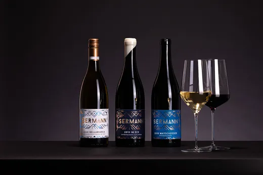Auf einer schwarzen Fläche sind drei Flaschen Sermann-Wein mit dekorativen Etiketten zu sehen, daneben zwei gefüllte Weingläser – eines mit Weißwein und eines mit Rotwein – vor einem dunklen Hintergrund.