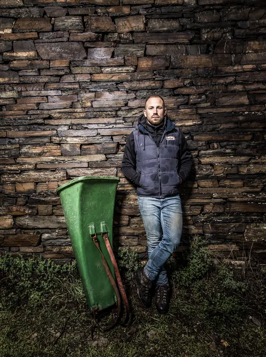 Ein Mann in Jeans und einer blauen Weste lehnt mit Armen in den Jackentaschen an einer Steinmauer und steht neben einem alten grünen Lesegerät auf einer Rasenfläche.