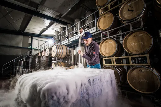 Eine Person rührt in einem Weingut in einem großen Behälter um, aus dem dichter weißer Dampf aufsteigt; um sie herum stehen Holzfässer auf Gestellen in einer Industriehalle.