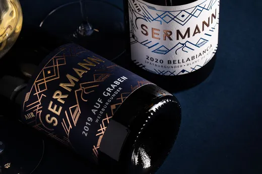 Auf einer dunkelblauen Fläche liegen zwei Weinflaschen mit der Aufschrift „Sermann“. Auf einer Flasche steht „2019 Auf Graben“ auf einem dunklen Etikett, auf der anderen „2020 Bellabianca“ auf einem weißen Etikett. Daneben ist teilweise ein Glas Weißwein zu sehen.