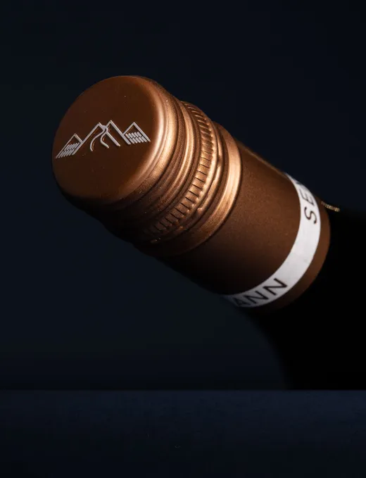 Nahaufnahme eines bronzefarbenen Schraubverschlusses für eine Weinflasche mit einem weißen Berg-Logo vor einem dunklen Hintergrund. 