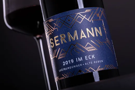 Eine Nahaufnahme einer Weinflasche mit der Aufschrift „SERMANN 2019 IM ECK SPÄTBURGUNDER • ALTE REBEN“ und goldenen geometrischen Mustern auf dunkelblauem Hintergrund, daneben ein teilweise sichtbares Glas Rotwein.
