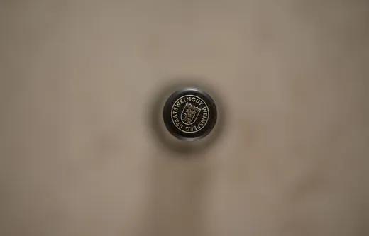Eine Nahaufnahme einer Weinkapsel aus der Vogelperspektive mit der Aufschrift „Staatsweingut weinberg“, vor einem unscharfen, neutralen Hintergrund. Die Oberseite der Kapsel in der Bildmitte ist scharf abgebildet.