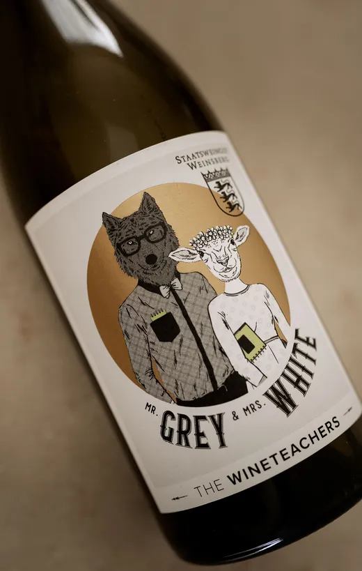 Eine Weinflasche mit einem Etikett, auf dem ein Wolf in einem karierten Hemd mit der Aufschrift „Mr. Grey“ und ein Schaf in einem Kleid mit der Aufschrift „Mrs. White“ abgebildet sind. Auf dem Etikett steht „THE WINETEACHERS“ und es hat einen goldenen Kreis als Hintergrund.