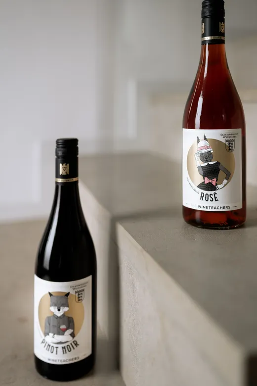 Zwei Weinflaschen auf einer Steintreppe. Die linke Flasche trägt die Aufschrift „Pinot Noir“ und ist mit der Abbildung eines wolfes mit Brille und Fliege verziert. Die rechte Flasche trägt die Aufschrift „Rosé“ und zeigt eine ähnliche Tieresabbildung.