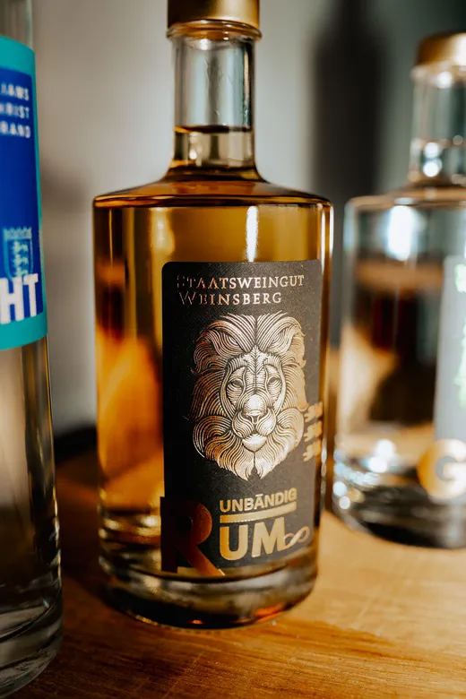 Eine Glasflasche Rum mit einem dunklen Etikett, auf dem eine detailreiche Löwenillustration sowie die Aufschriften „Staatsweingut Weinsberg“ und „Unbändig Rum“ zu sehen sind, steht zwischen zwei anderen Flaschen auf einer Holzunterlage.