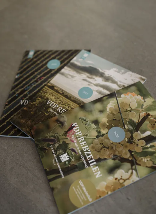 Drei Zeitschriften mit Naturmotiven auf dem Titelblatt – darunter Weinreben und Landschaften – liegen auf einer grauen Fläche ausgebreitet. Auf der obersten Zeitschrift sind weiße Weintrauben abgebildet, und darauf befindet sich ein rundes Etikett mit der Aufschrift „2022“.