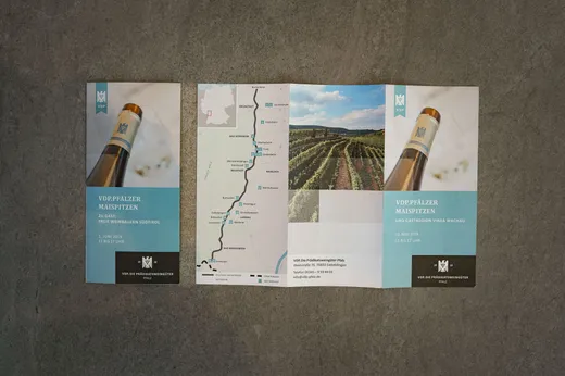 Eine dreifach gefaltete Broschüre liegt offen auf einer grauen Fläche. Die Broschüre zeigt eine Weinflasche, einen Weinberg, eine Routenkarte im Mittelteil, Veranstaltungsinformationen sowie einen deutschen Text, der für eine Weinverkostung wirbt.