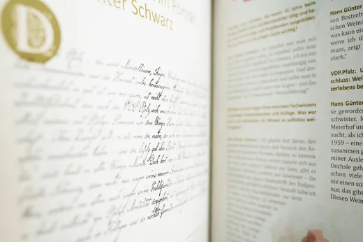 Eine Nahaufnahme eines aufgeschlagenen Buches zeigt eine Seite mit handschriftlichem Text in Schreibschrift auf der linken Seite und gedrucktem, maschinengeschriebenem Text auf der rechten Seite, beides auf Deutsch. Das Bild ist hell ausgeleuchtet und auf die handschriftliche Seite fokussiert.