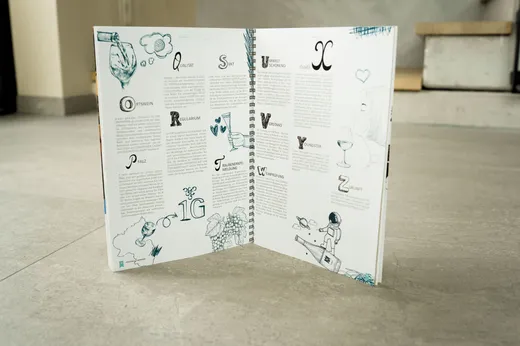 Ein offenes, spiralgebundenes Buch steht aufrecht auf einem hellgrauen Boden und zeigt illustrierte Seiten mit dekorativen Buchstaben, Zeichnungen und Text auf Deutsch. Im Hintergrund sind eine Treppe und eine beige Wand zu sehen.