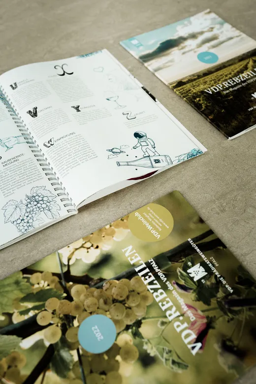 Auf einer beigen Fläche liegen drei Zeitschriften. Eine Zeitschrift ist aufgeschlagen und zeigt illustrierte Seiten, während die beiden anderen ihre farbenfrohen Titelseiten mit Weinbergen und Weintrauben präsentieren.