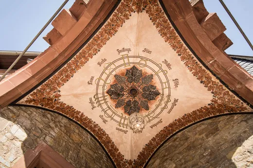 Eine verzierte Gewölbedecke mit einem kunstvollen kreisförmigen Motiv und deutschen Wörtern, die mit Blumenmustern verziert ist und von Steinmauern sowie rotbraunen architektonischen Elementen umgeben ist.