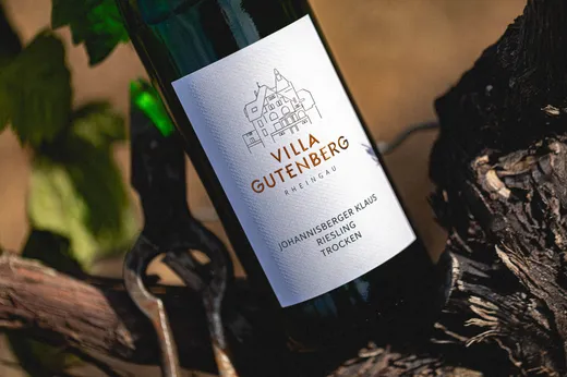 Eine Flasche „Villa Gutenberg Riesling“ steht auf einer Weinrebe; auf dem weißen Etikett ist eine Strichzeichnung eines Gebäudes zu sehen sowie der Text: „Villa Gutenberg Rheingau Johannisberger Klaus Riesling Trocken“.