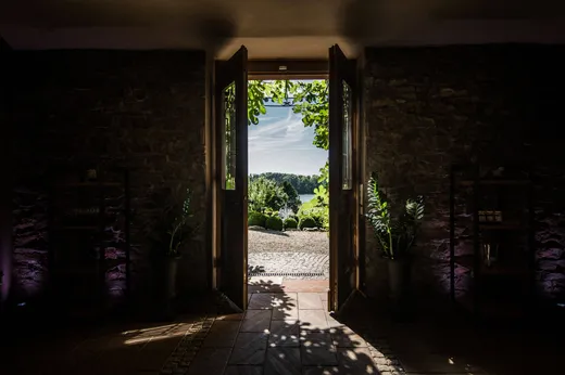 Blick durch die offenen Flügeltüren eines Raums mit Steinwänden auf einen sonnendurchfluteten Garten, mit üppig blühenden Pflanzen zu beiden Seiten und Sonnenlicht, das Schatten auf den Fliesenboden wirft. Draußen sind Bäume, der Himmel und viel Grün zu sehen.