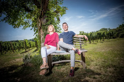 Zwei Menschen sitzen auf einer Holzbank unter einem Baum in einem Weinberg, lächelnd und entspannt. Es ist ein sonniger Tag mit blauem Himmel und grünen Weinreben im Hintergrund.