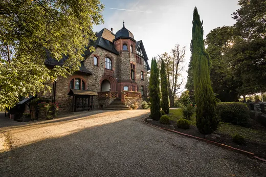 Am Ende einer geschwungenen Kiesauffahrt steht ein großes Steinhaus mit einem Turm, Bogenfenstern und einem steilen Dach, umgeben von hohen Bäumen und sorgfältig geschnittenen Sträuchern an einem sonnigen Tag.