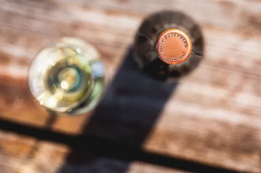 Eine Nahaufnahme von oben auf eine Weinflasche mit kupferfarbenem Verschluss neben einem unscharfen Glas Weißwein auf einer Holzfläche, das im Sonnenlicht einen Schatten wirft.