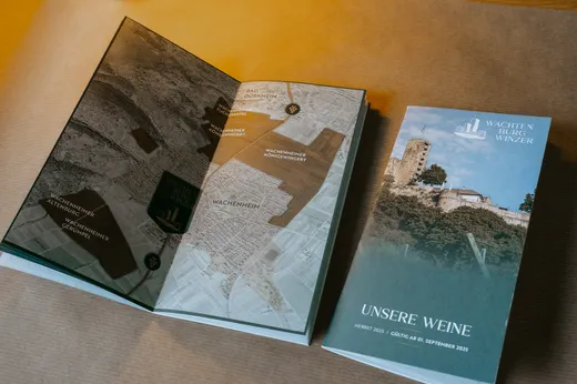 Eine Broschüre für Wachtenburg Winzer mit einem Titelblatt, auf dem ein Foto des Schlosses und der Text „UNSERE WEINE“ zu sehen sind, sowie einer aufgeschlagenen Seite, die eine Weinbergkarte mit verschiedenen beschrifteten Regionen zeigt.