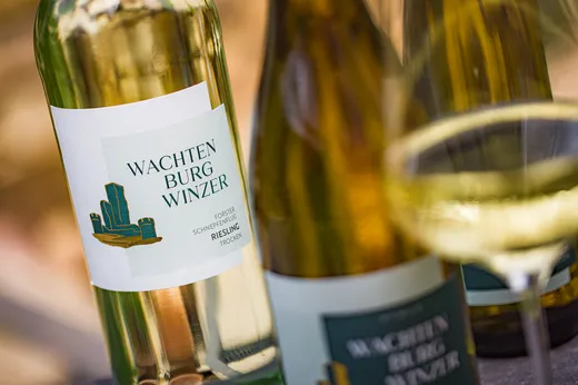 Eine Nahaufnahme einer Flasche mit der Aufschrift „Wachtenburg Winzer Riesling“ neben zwei weiteren Weinflaschen und einem gefüllten Weinglas, vor einem sanften Außenhintergrund.