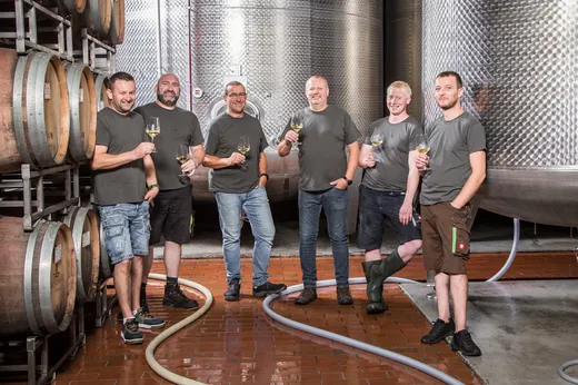 Sechs lächelnde Männer in Freizeitkleidung stehen gemeinsam in einem Weingut und halten Gläser mit Weißwein in der Hand. Sie sind auf einem Fliesenboden von Holzfässern und großen Metallgärtanks umgeben.