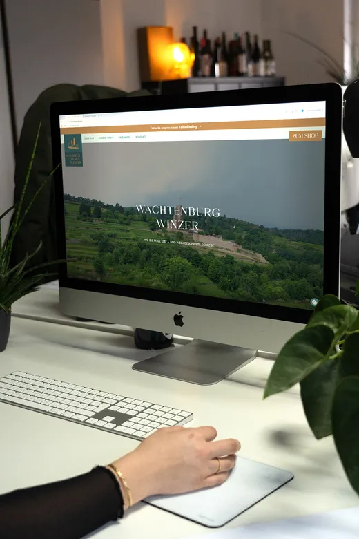 Eine Person nutzt einen Desktop-Computer, auf dem eine Website der „Wachtenburg Winzer“ mit einem malerischen Weinbergfoto angezeigt wird. Auf dem Schreibtisch stehen eine Tastatur, eine Maus und Grünpflanzen, im Hintergrund sind Weinflaschen zu sehen.