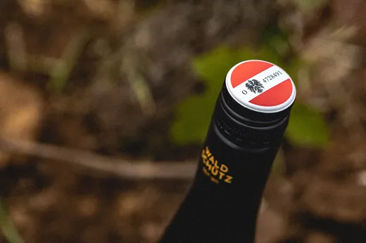 Eine Nahaufnahme des Verschlusses einer Weinflasche, auf dem ein rot-weißes Siegel mit einem schwarzen Adler-Emblem zu sehen ist. Der Hintergrund ist unscharf und weist Erdtöne sowie grünes Laub auf.