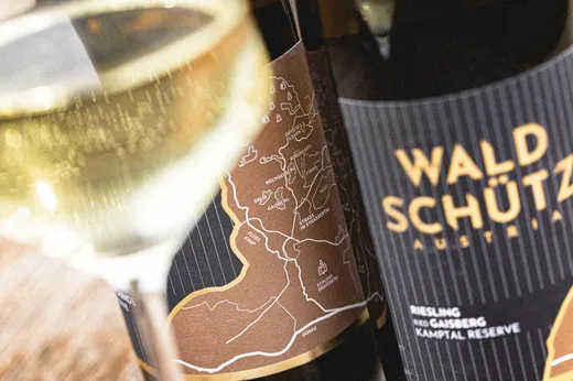 Eine Nahaufnahme eines mit Weißwein gefüllten Weinglases neben zwei Flaschen mit der Aufschrift „Waldschütz Austria Riesling“, deren Etiketten ein Kartendesign zeigen und die im Hintergrund leicht unscharf zu sehen sind.