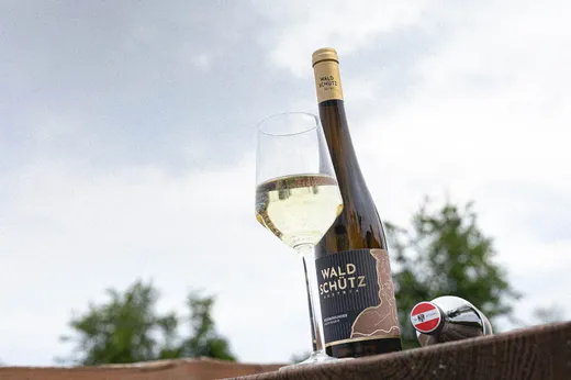 Zwei Flaschen Waldschütz-Wein und ein gefülltes Weinglas stehen im Freien auf einer Holzfläche, im Hintergrund sind grüne Bäume und ein bewölkter Himmel zu sehen.