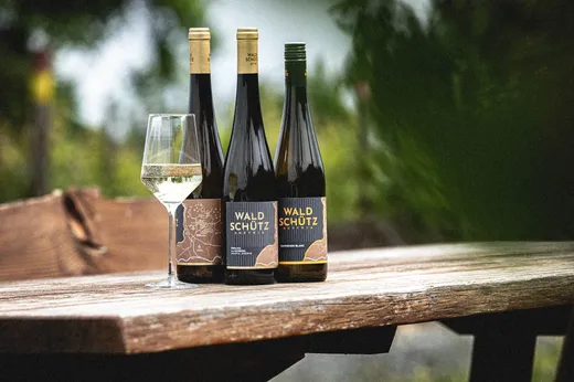 Drei Flaschen „Waldschütz“-Weißwein und ein gefülltes Weinglas stehen auf einem rustikalen Holztisch im Freien, im Hintergrund ist verschwommenes Grün zu sehen.