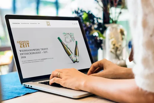 Eine Person tippt auf einem Laptop, auf dessen Bildschirm eine Website mit zwei Weinflaschen und einem deutschen Text über die Kunst der Weinherstellung zu sehen ist. Die Szene spielt an einem Tisch, im Hintergrund sind verschwommene Blumen und eine Vase zu sehen.