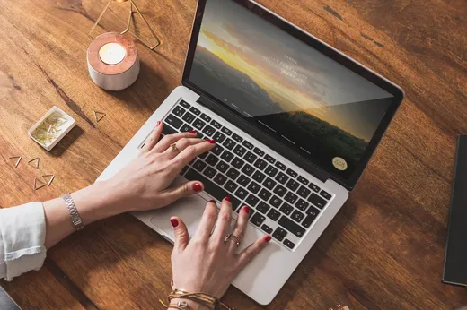 Eine Person mit rotem Nagellack sitzt an einem Holztisch und arbeitet an einem Laptop, umgeben von einer Kerze, goldenen Büroklammern, einem Armband und einem kleinen schwarzen Notizbuch. Auf dem Laptop-Bildschirm ist eine Website mit Landschaftsbildern zu sehen.
