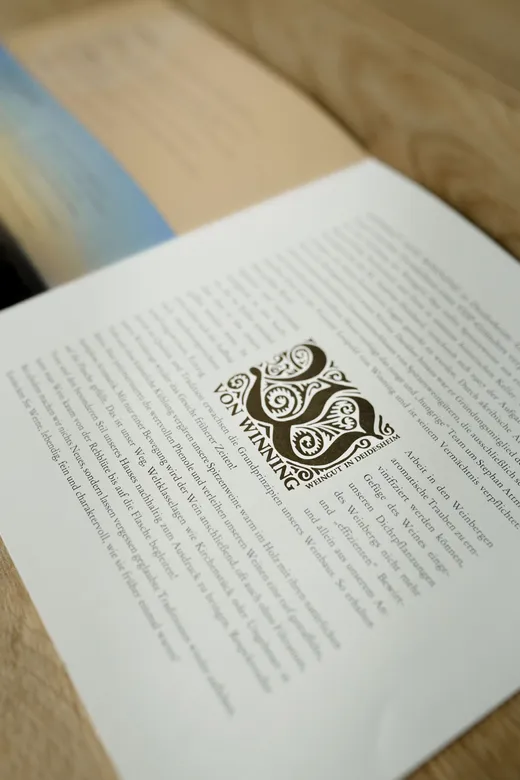 Eine Nahaufnahme eines aufgeschlagenen Heftes, das Textabschnitte auf Deutsch zeigt, mit einem dekorativen Logo und dem Schriftzug „VON WINNING“ in der Mitte der Seite. Das Heft liegt auf einer Holzunterlage.
