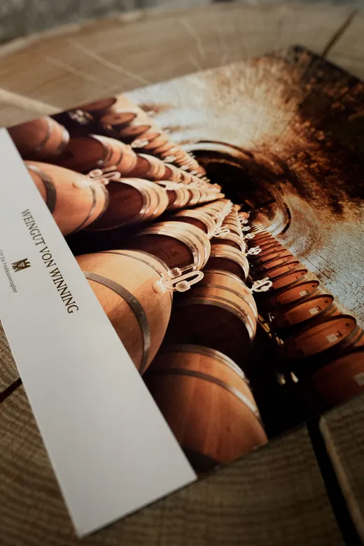 Eine Nahaufnahme einer Broschüre oder Postkarte mit einem Foto von Reihen hölzerner Weinfässer in einem schwach beleuchteten Weinkeller. Die Karte liegt auf einer hölzernen Unterlage, wobei der Text „WEINGUT VON WINNING“ zu sehen ist.