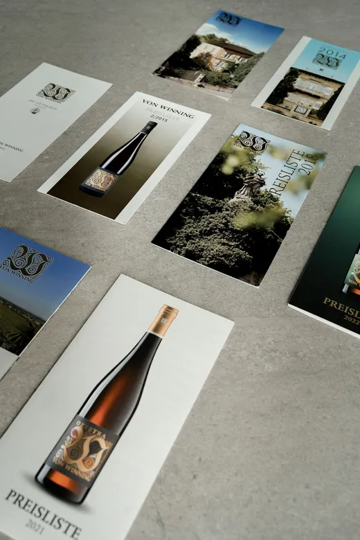 Auf einer grauen Fläche sind mehrere Weinpreislisten und Broschüren mit unterschiedlichen Cover-Motiven angeordnet, darunter Abbildungen von Weinflaschen, Weinbergen und Gebäuden. Auf einigen Covern ist der Schriftzug „PREISLISTE“ zu sehen.
