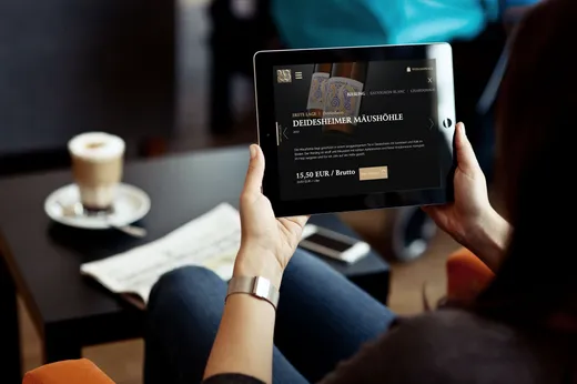 Eine Person, die ein Tablet auf dem Schoß hält und auf einer Website surft, im Hintergrund stehen auf dem Tisch ein Latte und eine Zeitung. Die Person sitzt auf einem Stuhl, trägt Jeans und eine Armbanduhr.