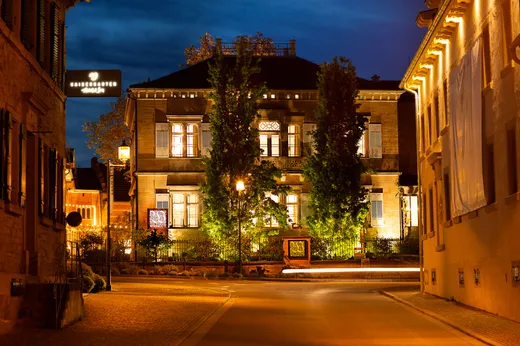 Ein warm beleuchtetes historisches Gebäude mit hohen Bäumen davor ist nachts in einer ruhigen Kopfsteinpflasterstraße zu sehen, umgeben von anderen beleuchteten Gebäuden.
