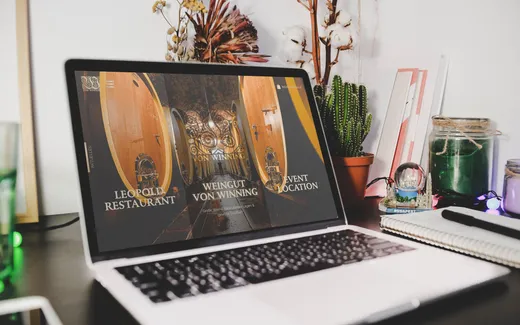 Auf einem Schreibtisch steht ein Laptop, auf dessen Bildschirm eine Website mit Abbildungen von Weinfässern aus Holz und einem Text über ein Restaurant, ein Weingut und einen Veranstaltungsort zu sehen ist. Auf dem Schreibtisch stehen Pflanzen, Gläser, Schreibutensilien, ein Notizbuch und eine Kristallkugel.