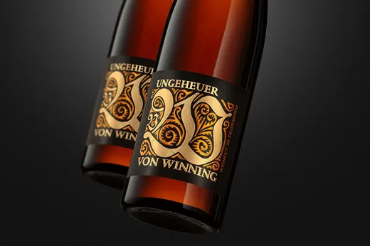 Zwei bernsteinfarbene Weinflaschen mit kunstvoll gestalteten schwarz-goldenen Etiketten, auf denen in eleganter Schrift „WEINGUT VON WINNING“ vor einem dunklen Hintergrund zu lesen ist.