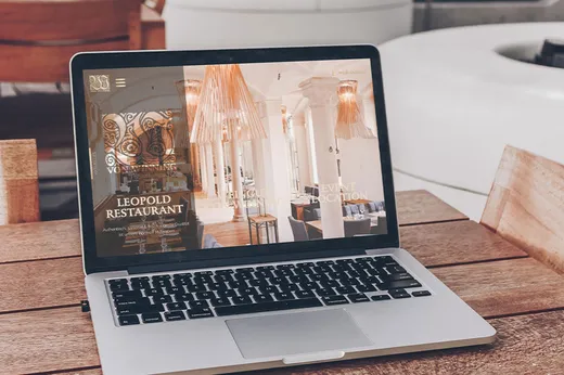 Auf einem Laptop, der auf einem Holztisch steht, ist die Website eines Restaurants zu sehen, auf der elegante Innenaufnahmen sowie Informationen zum Restaurant „Leopold“ und zu dessen Veranstaltungsdienstleistungen zu finden sind. Im Hintergrund sind weiße Stühle und ein Teil eines weiteren Tisches zu erkennen.
