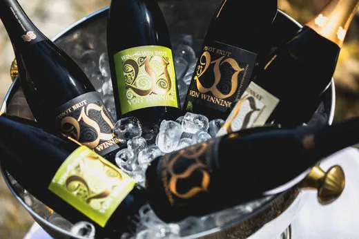Eine Nahaufnahme mehrerer Flaschen Von Winning-Sekt, die in einem Eisbehälter aus Metall gekühlt sind; die Etiketten in Grün, Gold und Schwarz sind von Eiswürfeln umgeben.